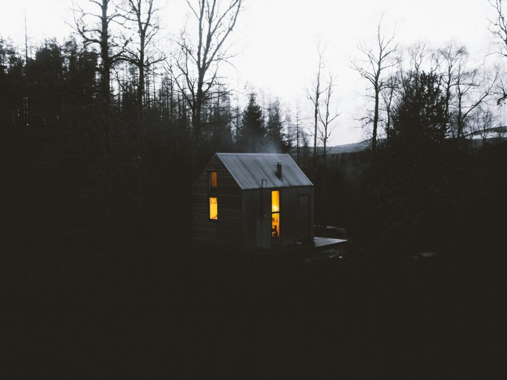 Cozy cabin in dark woods with glowing windows, chimney smoke. Rustic retreat, peaceful escape, nature scene.